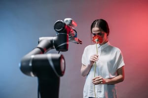 A robot arm gives a flower to a young lady who smells the flower's scent.